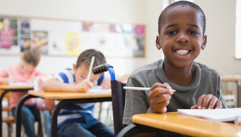 Descrição da imagem #pracegover: Em uma sala de aula, vemos três alunos, sentados em suas carteiras. Em primeiro plano, vemos um aluno cadeirante. Os alunos estão realizando uma atividade. O aluno em evidência segura um lápis com a mão direita, enquanto apoia a mão esquerda em um caderno, que está em cima da mesa. O menino cadeirante é negro, tem cabelos curtos e usa uma camiseta na cor cinza. Ele está sorrindo e olhando para a câmera. e Fim da descrição.