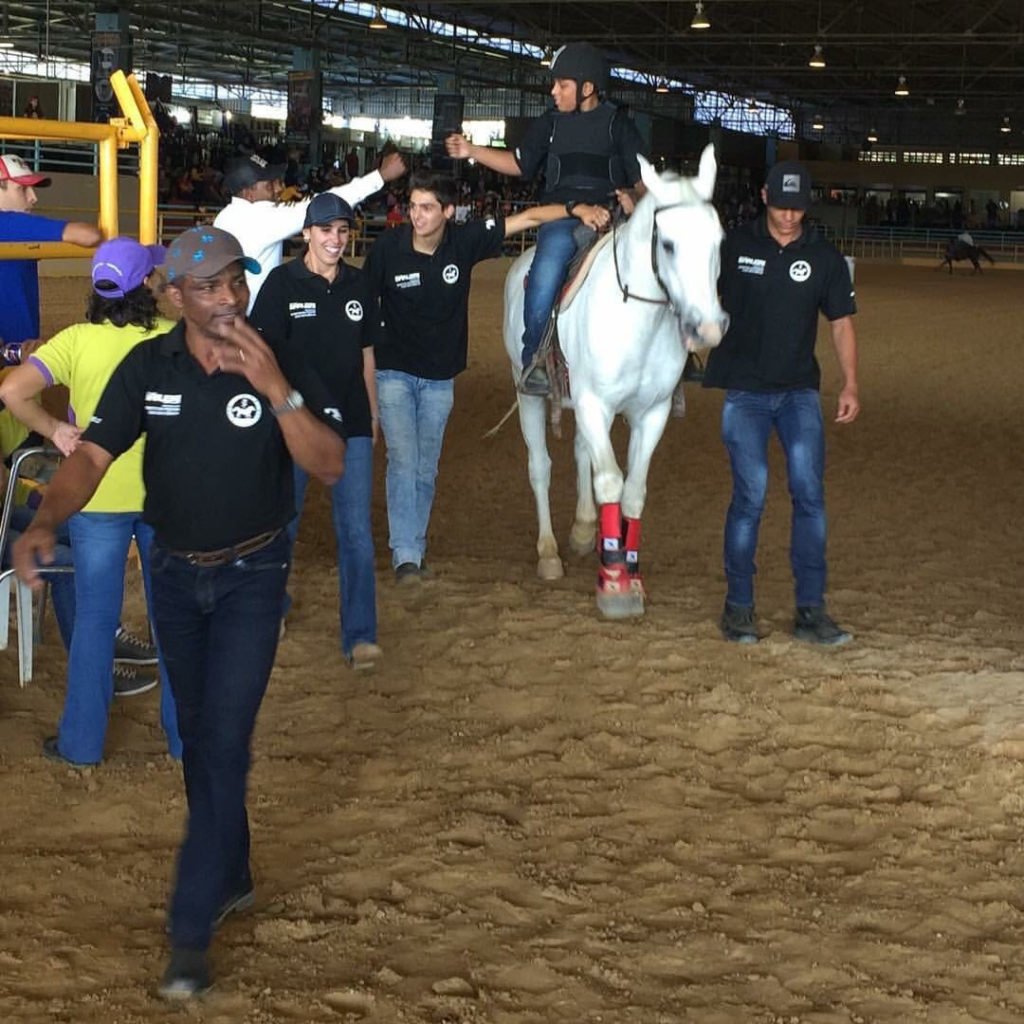 Descrição da imagem #PraCegoVer: Imagem no formato quadrado. Um grupo de profissionais da equoterapia acompanham um rapaz durante uma apresentação da prova dos 3 Tambores. O rapaz está montado em um cavalo branco. Fim da descrição.
