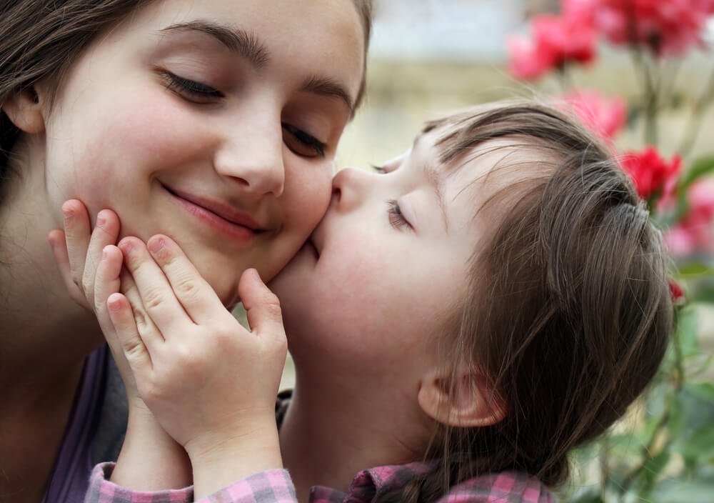 Descrição da imagem #pratodosveres: Duas meninas, uma maior e uma menor. A menina menor tem síndrome de Down. Ela está segurando o rosto da menina maior e dando um beijo em sua bochecha. As duas estão com os olhos fechados, e com expressão de felicidade. Ao fundo, vemos algumas flores vermelhas em um jardim. Fim da descrição.