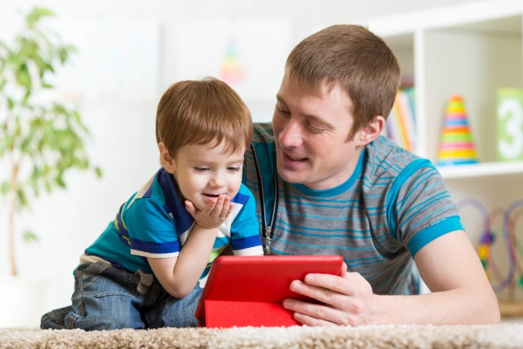 Descrição da imagem #PraCegoVer: Imagem no formato retangular, na horizontal. Um ambiente doméstico. Pai e filho estão lado a lado assistindo algo em um tablet. Eles estão vestidos com camiseta listrada, em tons predominantes de azul. Fim da descrição.