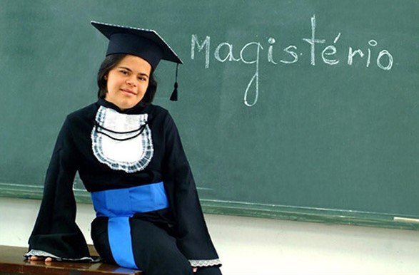 Descrição da imagem #PraCegoVer: A professora Débora Seabra em uma sala de aula. Ela está vestindo uma beca com faixa azul, em referência ao magistério. Débora é uma moça morena, com a pele clara, e cabelos pretos. Ela usa também um capelo na cabeça. Atrás dela tem uma lousa, onde está escrito a palavra Magistério, em giz branco. Fim da descrição.