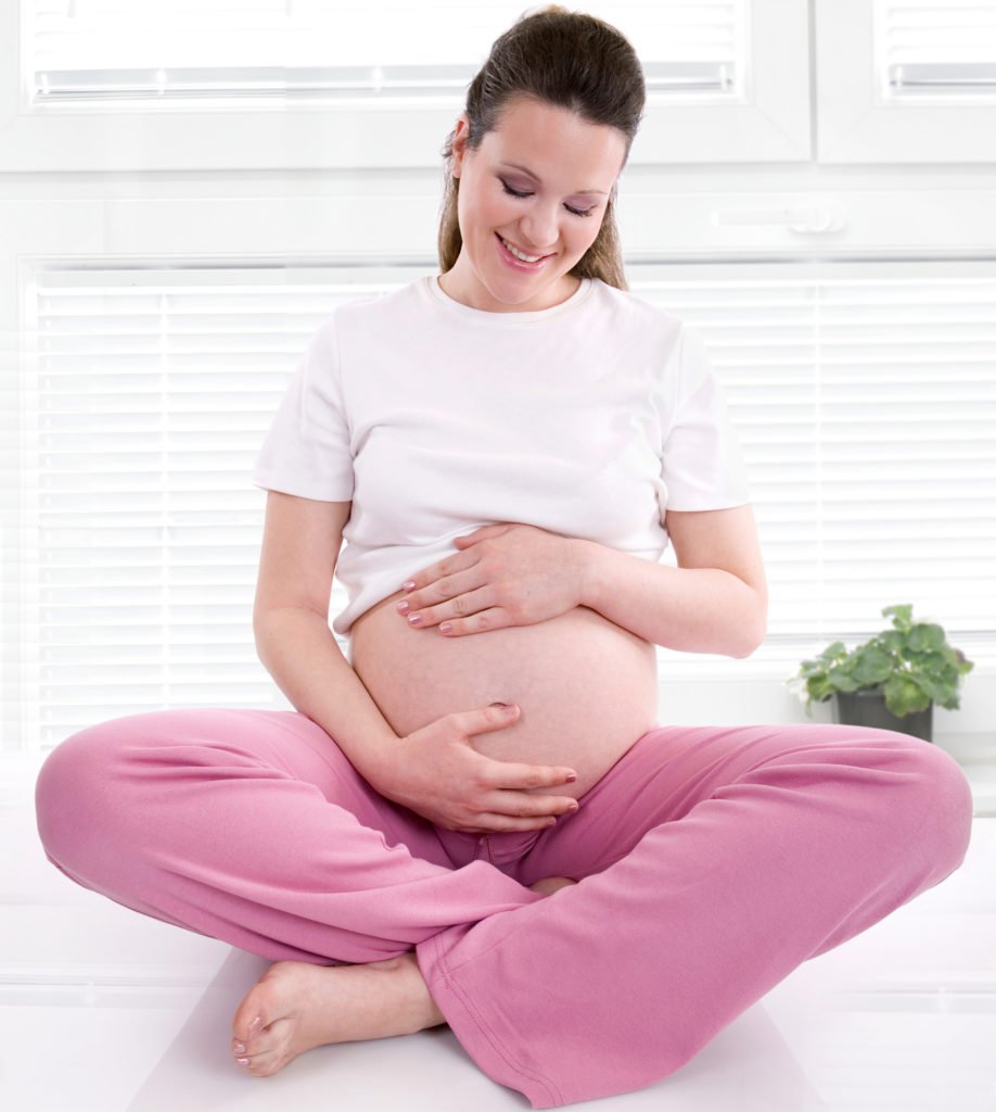 Descrição da imagem #PraCegoVer: Imagem no formato quadrado. Uma mulher grávida está sentada no chão. Ela tem as mãos em volta da barriga e está sorrindo. Fim da descrição.