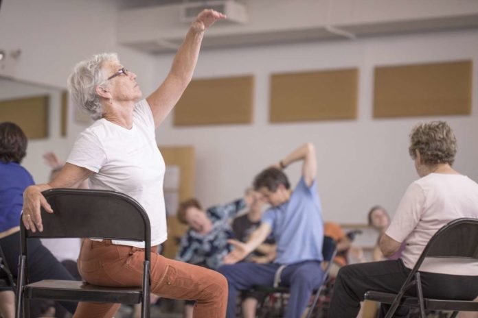 Descrição da imagem #PraCegoVer: Imagem no formato retangular, na horizontal. Cena do documentário 'Como se eu estivesse voando'. Uma sala, onde as pessoas realizam alguns movimentos, como uma coreografia. Em primeiro plano, vemos uma mulher idosa sentada de lado em uma cadeira. Ela tem o braço esquerdo esticado para cima, e o braço direito apoiado no encosto da cadeira. Fim da descrição.