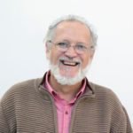 Descrição da imagem #PraCegoVer: Imagem no formato quadrado. João Felippe, está sorrindo. João é um homem que tem pele morena, cabelos e barbas brancas. Ele está usando uma camisa rosa e uma malha marrom. Fim da descrição.