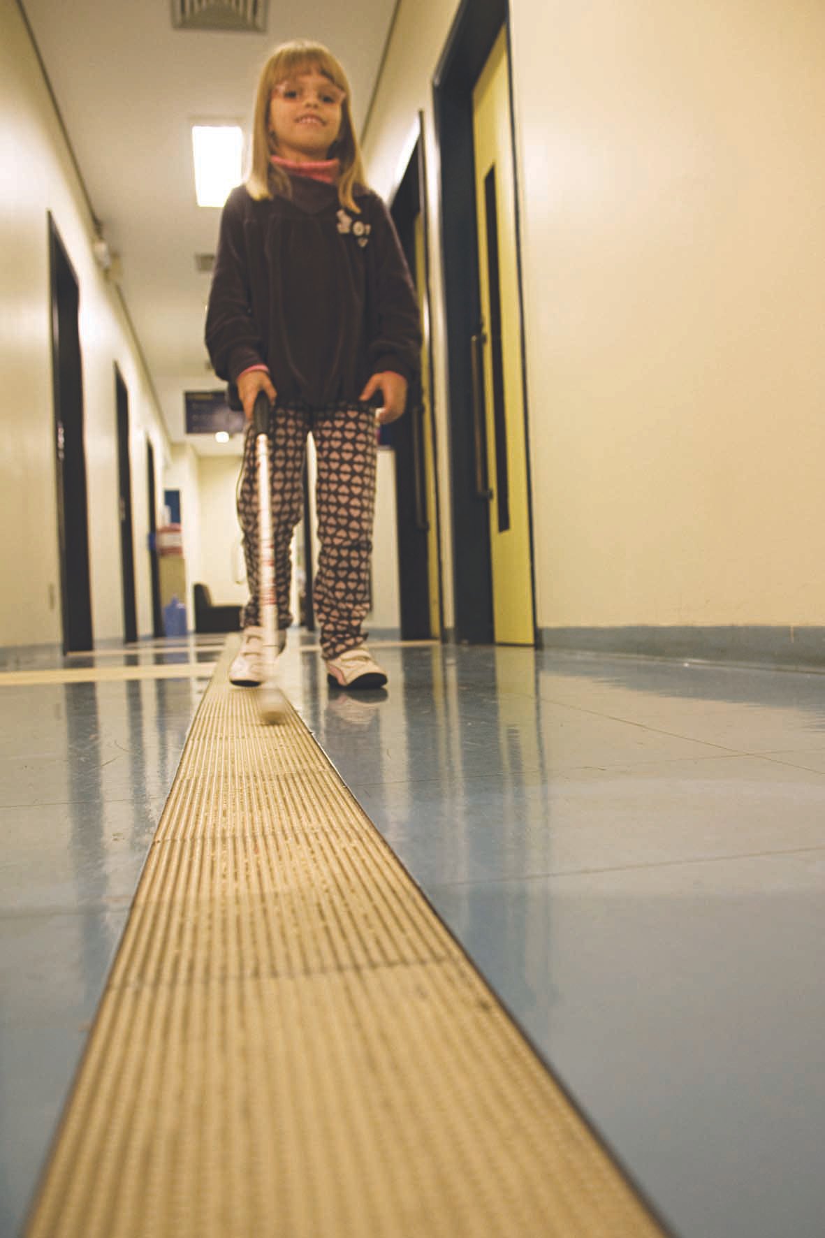 Descrição da imagem #PraCegoVer: Imagem no formato retangular, na vertical. Uma menina está no corredor da Laramara. Ela tem deficiência visual e está caminhando com a ajuda de uma bengala e do piso podotátil. Fim da descrição.