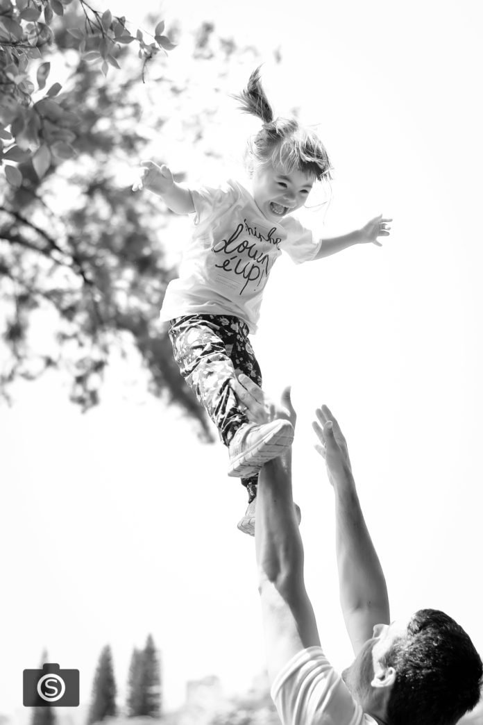 Descrição da imagem #PraCegoVer: Imagem no formato retangular, na vertical. Um homem joga uma menina que tem síndrome de Down para o alto. A menina se diverte com a brincadeira. Eles são pai e filha durante um dos eventos promovidos pelo projeto Minha Down é Up. A foto é preta e branca. Fim da descrição.