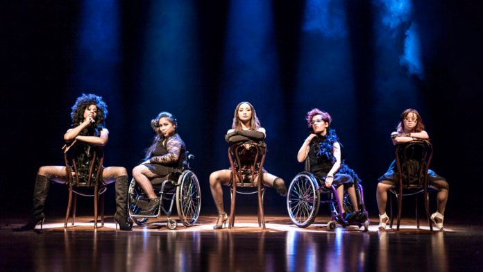 Descrição da imagem #pracegover: Foto de uma apresentação, que inspirada em uma cena do filme Chicago. Cinco mulheres estão no palco, sentadas em cadeiras, duas delas, em cadeiras de rodas. Fim da descrição.