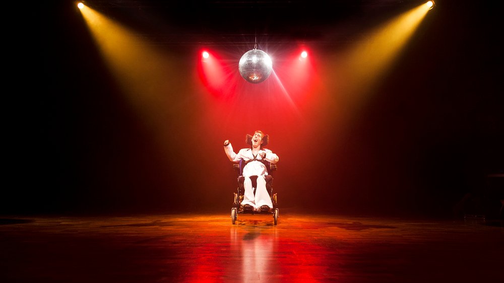 Descrição da imagem #PraCegoVer: Imagem no formato retangular, na horizontal. Cena inspirada no filme Os embalos de sábado à noite. Um rapaz que tem paralisia cerebral está sentado em sua cadeira de rodas, no centro do palco. Ele usa uma roupa típica dos anos 1970. Luzes coloridas dão o tom de discoteca à imagem. Fim da descrição.