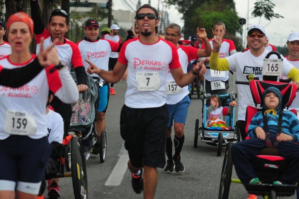 Descrição da imagem #PraCegoVer: Imagem no formato retangular, na horizontal. Um grupo de participantes do projeto Pernas de Aluguel durante uma corrida, que teve a participação de pessoas com deficiência. Entre os corredores, vemos três participantes em cadeiras de rodas. Fim da descrição.