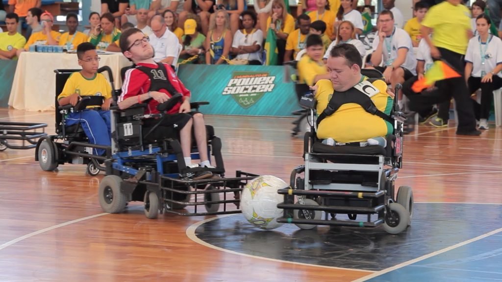Descrição da imagem #PraCegoVer: Imagem no formato retangular, na horizontal. Uma partida de Power Soccer. Na quadra vemos alguns atletas disputando a posse de bola. Em destaque, temos dois atletas. Um deles veste uma camisa vermelha e outro uma camisa amarela. Ao fundo é possível ver a torcida na arquibancada. Fim da descrição.