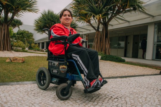 Descrição da imagem #PraCegoVer: Imagem no formato retangular, na horizontal. O colaborador Claudio Medica. Ele está em sua cadeira de rodas. Claudio é um rapaz moreno, de pele clara. Ele tem cabelos castanhos. Claudio usa uma cança preta e jaqueta vermelha. Fim da descrição.
