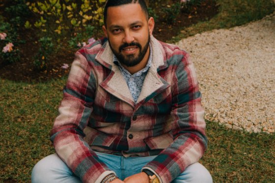 Descrição da imagem #PraCegoVer: Imagem no formato retangular, na horizontal. O colaborador Rafael. Ele está sentado no jardim da empresa. Rafael é um rapaz moreno, que tem cabelos castanhos e barba. Ele usa uma calça jeans e camisa xadrez. Fim da descrição.