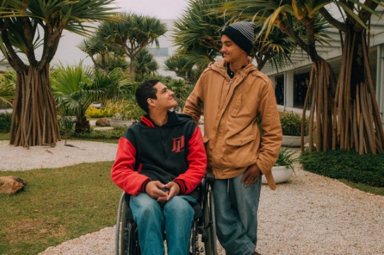 Descrição da imagem #PraCegoVer: Imagem no formato retangular, na horizontal. Os colaboradores Wesley e Gabriel, que são irmãos. Wesley está em sua cadeira de rodas e Gabriel está em pé, ao seu lado. Eles estão no jardim da empresa e se olham. Wesley e Gabriel são morenos. Wesley usa calça jeans e jaqueta preta e vermelha. Gabriel usa calça jeans e jaqueta bege. Fim da descrição.