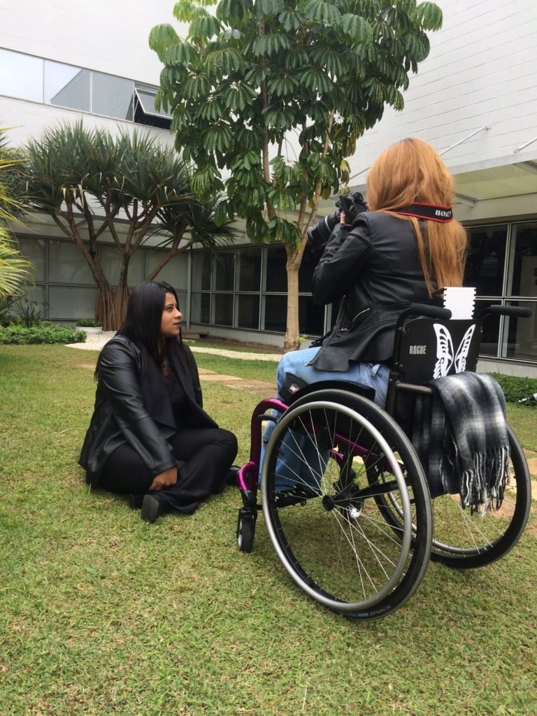 Descrição da imagem #PraCegoVer: Imagem no formato retangular, na vertical. A fotógrafa Maria Paula Vieira durante o ensaio fotográfico com os colaboradores com deficiência da TIM. A fotógrafa está sentada em sua cadeira de rodas, fotografando a colaboradora Jéssica, que sentada na grama. Fim da descrição.