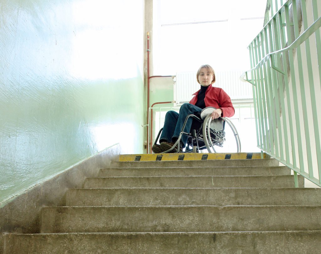 Descrição da imagem #PraCegoVer: Imagem no formato quadrado. Uma mulher está sentada em sua cadeira de rodas, na frente de uma escada. Fim da descrição.