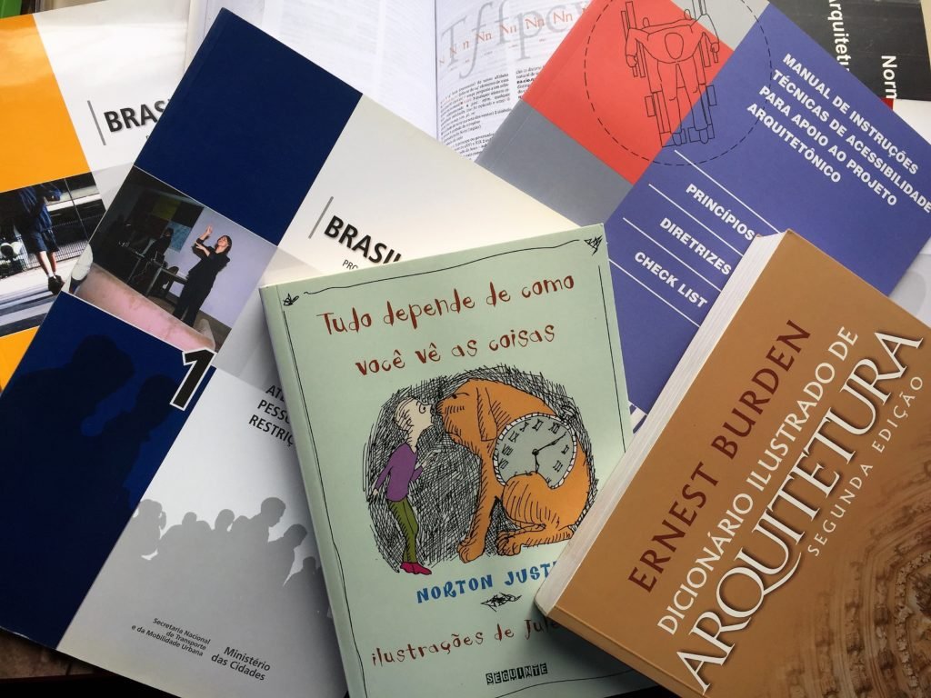Descrição da imagem #PracegoVer: Imagem no formato retangular, na horizontal. Alguns livros estão sobrepostos em cima de uma mesa. Fim da descrição.