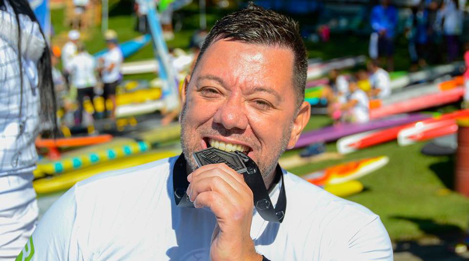 Descrição da imagem #PraCegoVer: Imagem no formato retangular, na horizontal. O atleta Alan Mazzoleni após conquistar uma medalha na modalidade. Alan está mordendo a medalha. Fim da descrição.