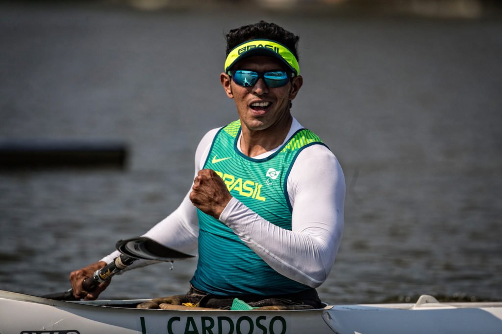 Descrição da imagem #PraCegoVer: Imagem no formato retangular, na horizontal. O atleta Luis Carlos Cardoso, que conquistou medalha de ouro no Mundial de Paracanoagem. Luis está em seu caiaque, durante o torneio. Ele é um homem moreno e está com uma regata do uniforme da delegação brasileira de paracanoagem. Luis usa uma camiseta branca, de mangas compridas, por baixo da regata. Ele está com uma viseira e óculos de sol. Fim da descrição.