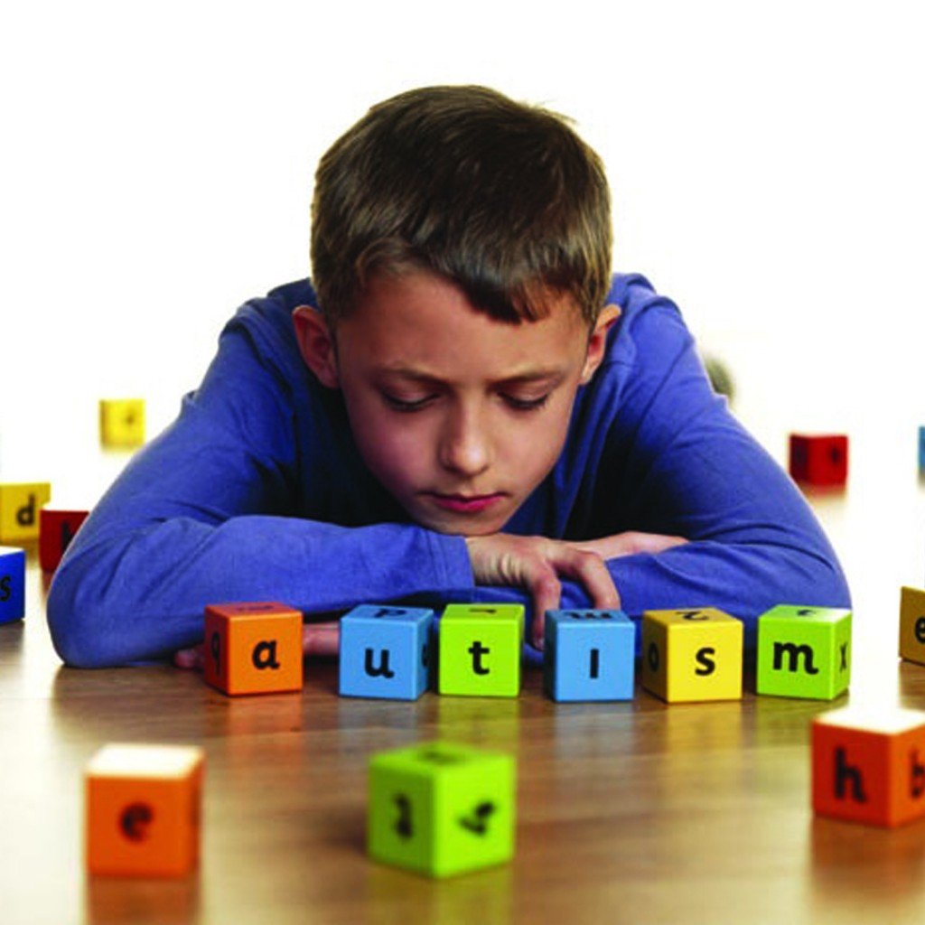 Descrição da imagem #PraCegoVer: Imagem no formato retangular, na horizontal. Um menino está sentado, em frente à uma mesa de madeira. Na mesa estão peças coloridas de blocos de montar, formando a palavra autism, autismo em português. O menino está com a cabeça apoiada nos braços, que estão cruzados sobre a mesa. Fim da descrição.