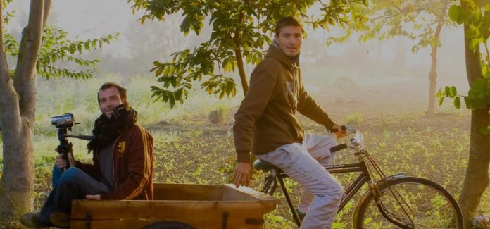 Descrição da imagem #PraCegoVer: Imagem no formato retangular, na horizontal. Dois rapazes estão em uma floresta. Um deles está sentado em um banco, segurando uma câmera, e o outro está em uma bicicleta. Fim da descrição.