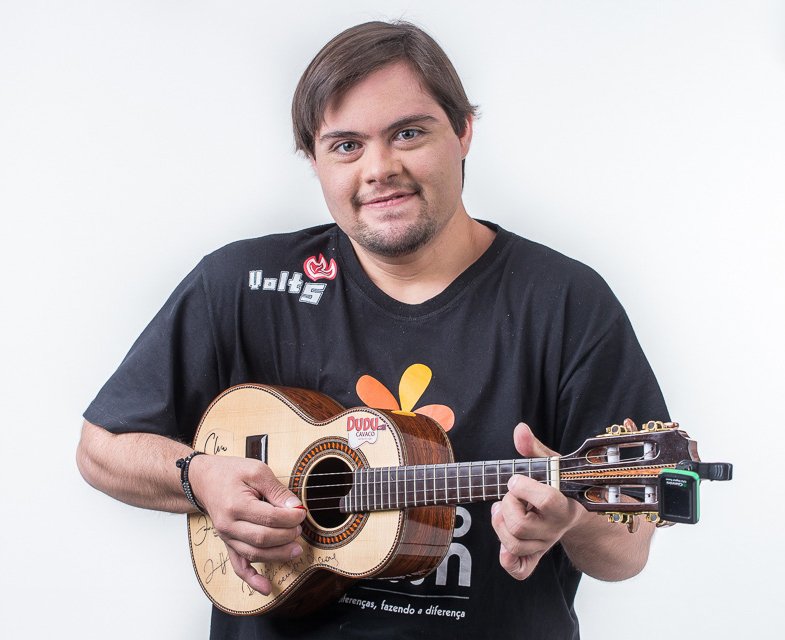 Descrição da imagem #pratodosverem: O músico Dudu do Cavaco. Ele está com uma camiseta preta, do Instituto Mano Down e segura seu cavaquinho. Dudu está sorrindo. Fim da descrição.
