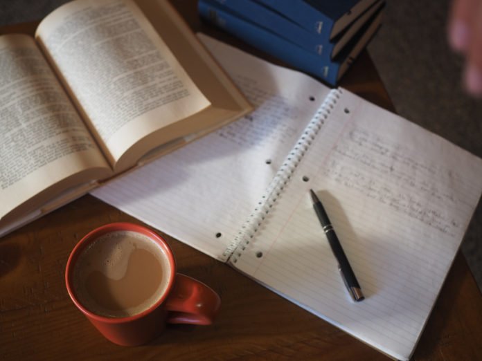 Descrição da imagem #PraCegoVer: Imagem no formato retangular, na horizontal. Em cima de uma mesa, um livro e um caderno abertos. Sobre o caderno está uma caneta preta e ao lado, está uma caneca com café e leite. Fim da descrição.