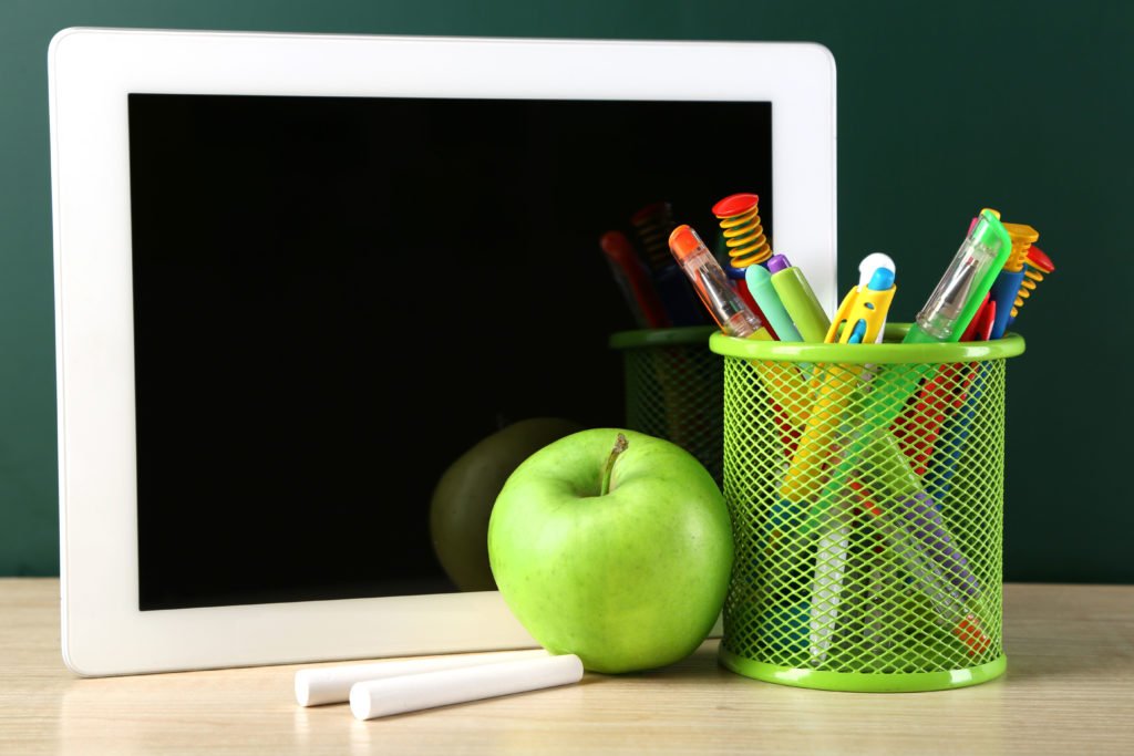 Descrição da imagem #PraCegoVer: Imagem no formato retangular, na horizontal. Sobre a mesa estão um tablet, um giz branco, uma maçã verde e um porta lápis verde, com lápis e canetas coloridas dentro. Fim da descrição.