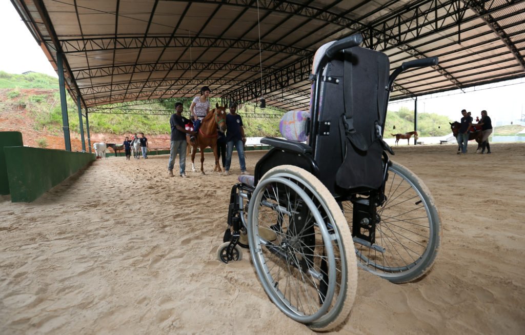 Descrição da imagem #PraCegoVer: Picadeiro da Hípica Santa Terezinha. Vemos, em primeiro plano, uma cadeira de rodas, e ao fundo, as pessoas durante a sessão no cavalo. Fim da descrição.