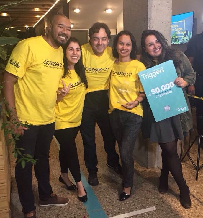 Descrição da Imagem #PraCegoVer: Foto no formato quadrado. Cinco pessoas, todas usando camiseta amarela. Da esquerda para direita: Jansey Oliveira (Portal Acesse), Patricia Senise (Hand Talk), Mauro Rezende (Youtube), Juliana Reis (Portal Acesse) e Bianca Ponte (Portal Acesse) segurando o cartão de crédito da Trigg ilustrativo em tamanho grande, com o valor de 50 mil reais. Fim da descrição.