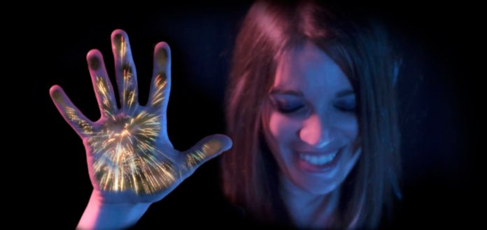 Descrição da imagem #PraCegoVer: Imagem no formato retangular, na horizontal. Uma mulher sentem os efeitos táteis dos fogos de artifício. Ela toca a tela, em que os efeitos estão sendo projetados. Fim da descrição.