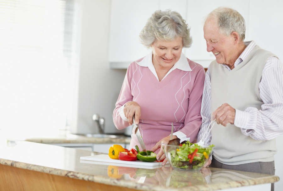Descrição da imagem #PraCegoVer: Imagem no formato retangular, na horizontal. Um casal de idosos está em uma cozinha preparando alguns alimentos. Fim da descrição.