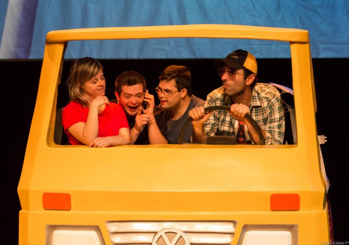 Descrição da imagem #PraCegoVer: Imagem no formato retangular, na horizontal. Cena da peça Colegas no Teatro. Os atores João Simões Junior, Ian Pereira e Giulia Merigo, que têm síndrome de Down, estão dentro de um carro cenográfico, amarelo, Eles estão muito animados. Fim da descrição.