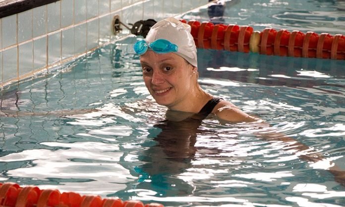 Descrição da imagem #PraCegoVer: Imagem no formato retangular, na horizontal. A atleta Kelly Antunes, que tem síndrome de Down, está dentro de uma piscina. Ela usa um maiô preto, toca branca na cabeça e óculos de natação azul. Kelly está sorrindo. Fim da descrição.