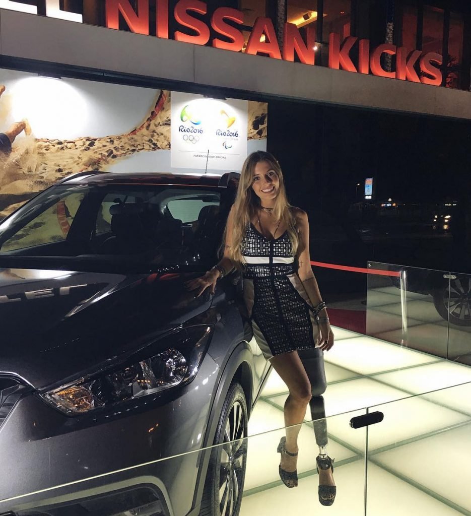 Descrição da imagem #PraCegoVer: Imagem no formato quadrado. A influenciadora digital Paola Antonini, que foi eleita embaixadora da Nissan para o público com deficiência. Paola é uma mulher jovem, tem cabelos loiros, lisos e compridos. Ela teve a perna esquerda amputada e usa uma prótese. Ela está encostada em um carro, modelo Nissan Kicks. Ao fundo vemos o letreiro com o nome do carro. Fim da descrição.