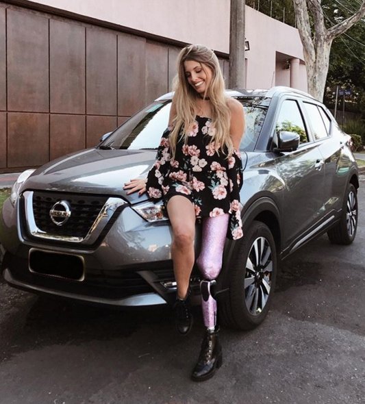 Descrição da imagem #PraCegoVer: Imagem no formato quadrado. A influenciadora digital Paola Antonini, que foi eleita embaixadora da Nissan para o público com deficiência. Paola é uma mulher jovem, tem cabelos loiros, lisos e compridos. Ela teve a perna esquerda amputada e usa uma prótese cor de rosa. Ela está encostada em um carro, modelo Nissan Kicks, e usa uma . Ela está usando um vestido preto com estampa florida, em tons de rosa. Fim da descrição.
