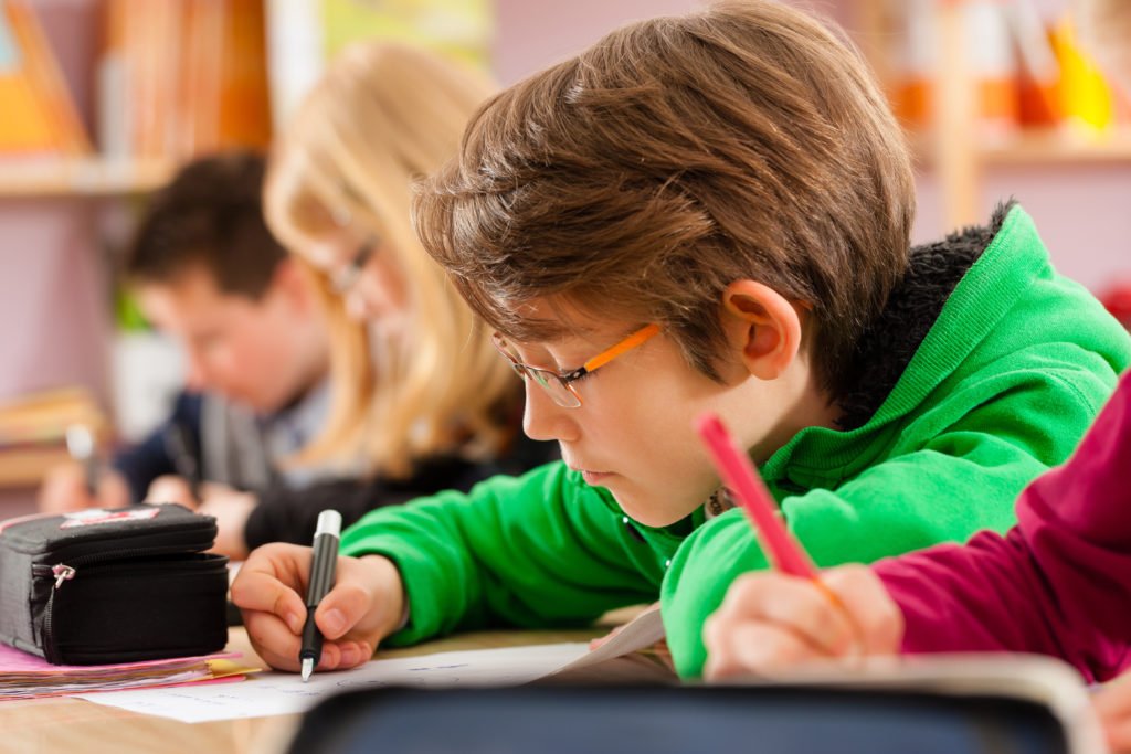 Descrição da imagem #PraCegoVer: A imagem está no formato retangular, na horizontal. Quatro crianças estão em uma sala de aula, sentadas lado a lado. Elas estão escrevendo. Fim da descrição.