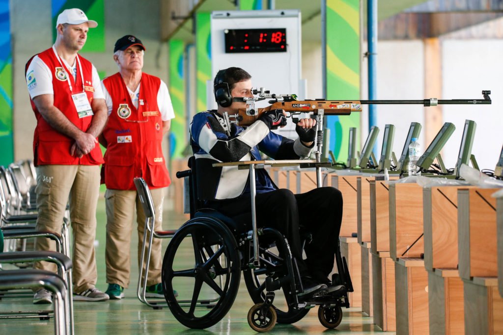 Descrição da imagem #PraCegoVer: Imagem no formato retangular, na horizontal. O paratleta Alexandre Galgani, durante torneio de Tiro Esportivo. Ele está sentado em sua cadeira de rodas, e utiliza seu rifle que está apoiado em um suporte especial. Fim da descrição.