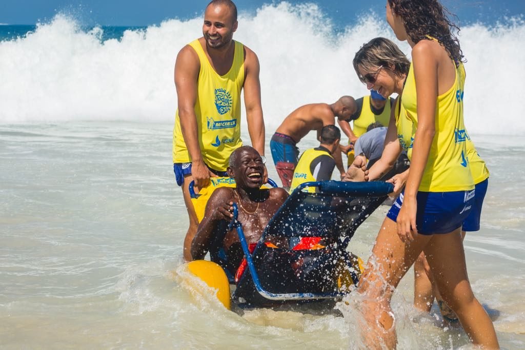 Descrição da imagem #PraTodosVerem: Imagem no formato retangular, na horizontal. Um homem está sentado na cadeira anfíbia, dentro do mar. Ele está sendo amparado por dois jovens que seguram a cadeira e uma outra pessoa está à frente, supervisionando o banho de mar. Fim da descrição.