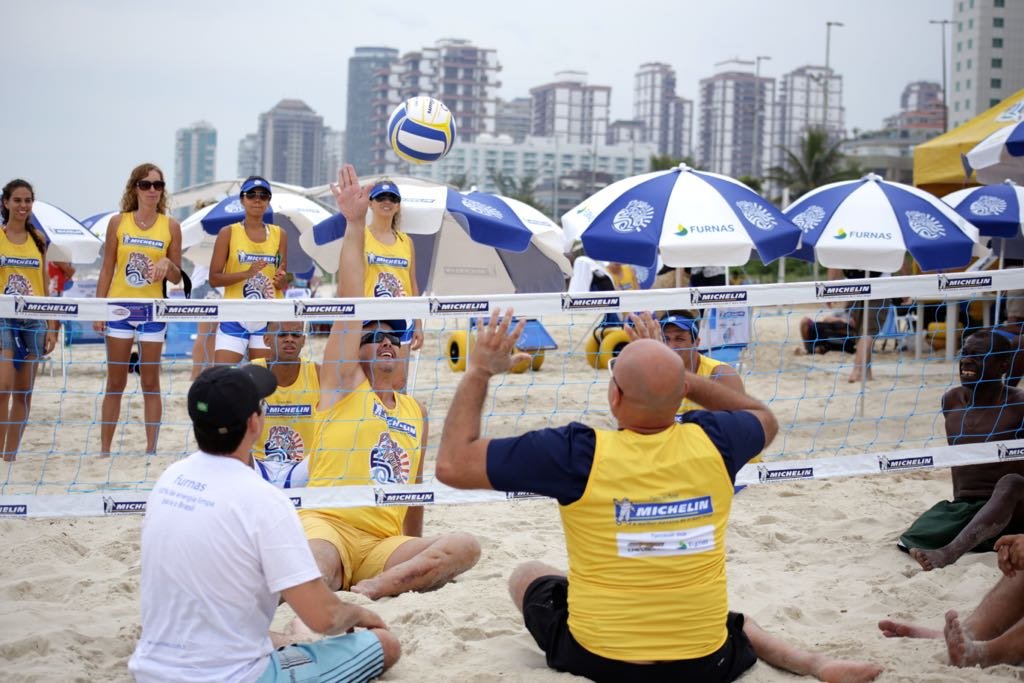 Descrição da imagem #PraTodosVerem: Imagem no formato retangular, na horizontal. Um grupo de pessoas está sentado na areia, jogando vôlei adaptado. Fim da descrição.