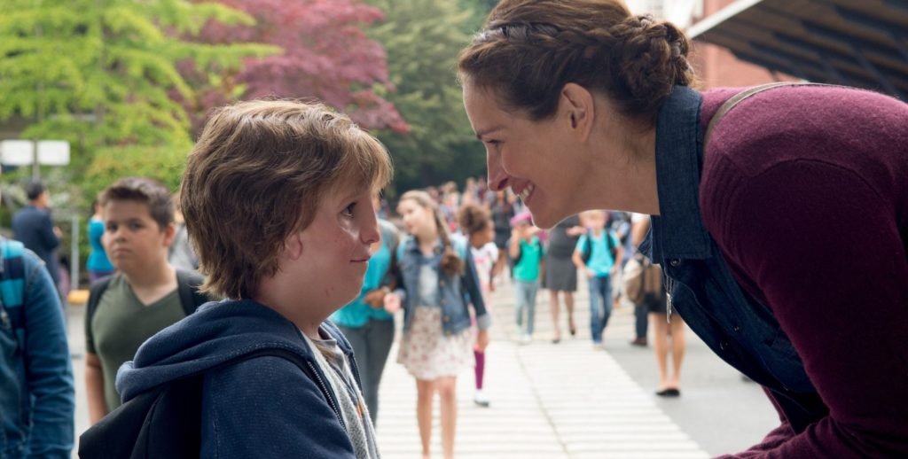 Descrição da imagem #PraCegoVer: Imagem no formato retangular, na horizontal. Cena do filme Extraordinário. Nela Julia Roberts conversa com o menino, protagonista da história, na porta da escola. Eles estão de perfil, e ao fundo vemos uma movimentação de pessoas. O menino tem uma malformação facial. Fim da descrição.