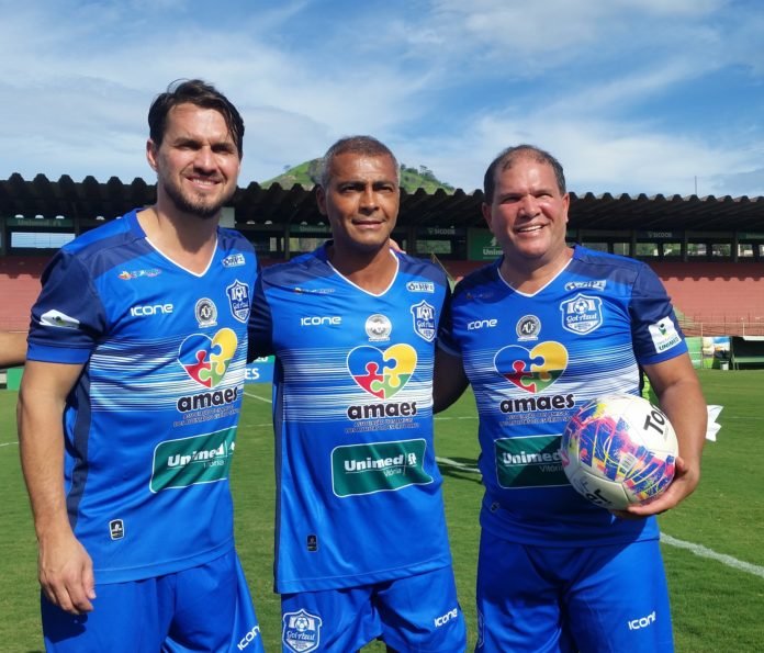 Descrição da imagem #PraCegoVer: Imagem no formato quadrado. Nela estão os ex-jogadores de futebol: Sávio, Romário e Geovani Silva. Eles estão em um estádio de futebol e usam o uniforme do evento Gol Azul. Geovani segura uma bola de futebol. Ao fundo vemos as arquibancadas e um lindo céu azul, além de parte do gramado verde. Fim da descrição.