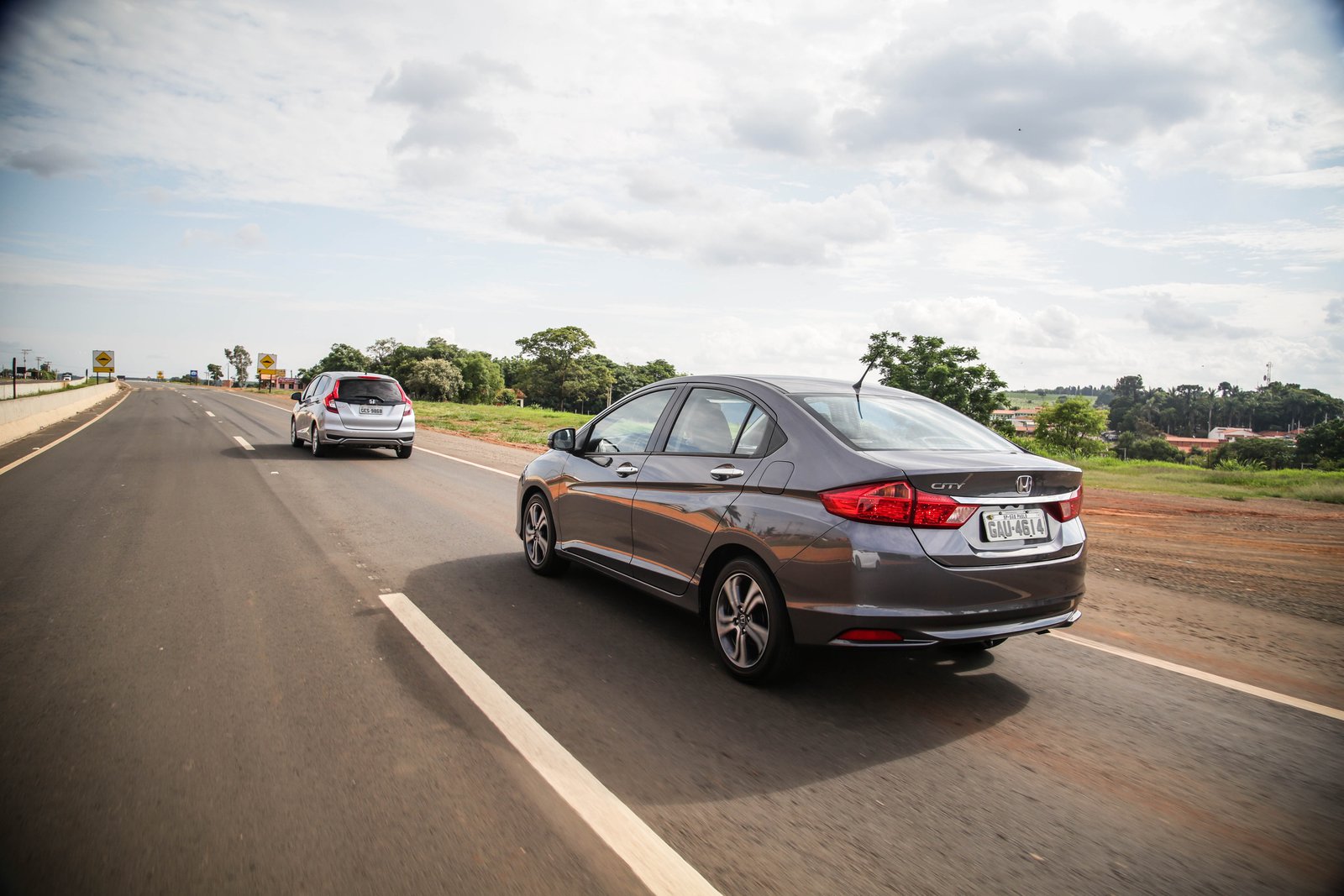 Descrição da imagem #PraCegoVer: A imagem está no formato retangular, na horizontal. Nela vemos dois modelos da Honda: um Fit e um City. Os carros estão na estrada, durante o test-drive. Fim da descrição.