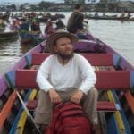 Descrição da imagem #PraTodosVerem: Tony Giles durante viagem a Indonésia. Ele está sentado em um pequeno barco. Fim da descrição.