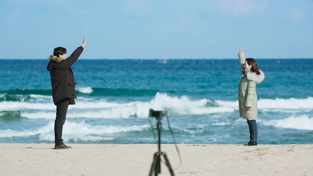Descrição da imagem #PraTodosVerem: Imagem no formato retangular, na horizontal. Os atores do curta-metragem estão em uma praia. Ao fundo vemos o mar, e ao centro, entre eles, está uma câmera fotográfica, em um tripé. Fim da descrição.