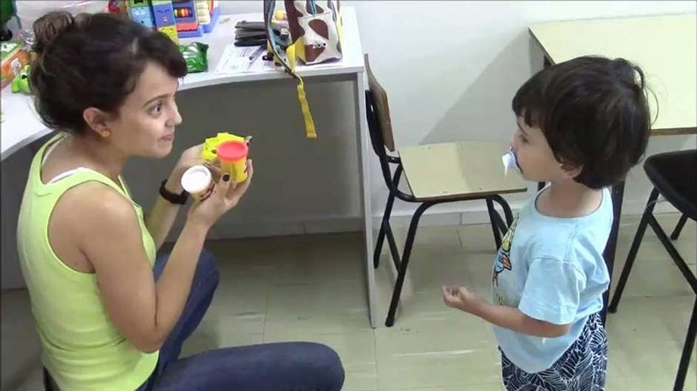 Descrição da imagem #PraTodosVerem: Imagem no formato retangular, na horizontal. Uma profissional está atendendo uma criança com autismo. Ela está abaixada em frente de criança, e interage com ele, usando um objeto colorido. Fim da descrição.