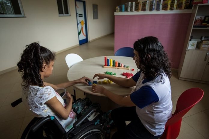 Descrição da imagem #PraTodosVerem: Imagem no formato retangular, na horizontal. Uma jovem, com deficiência física, está sentada em sua cadeira de rodas, ao lado de uma mulher. Elas estão em frente a uma mesa e realizam atividades com algumas peças coloridas. Fim da descrição.