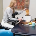 Descrição da imagem #PraTodosVerem: Imagem no formato retangular, na vertical. Uma mulher está sentada na cadeira de rodas robótica. Ela tem a parte da frente do tronco apoiado na cadeira, e está sentada em frente a uma mesa, comendo. A cadeira tem estrutura azul, rodas e acessórios pretos, além de formato oval. Fim da descrição.