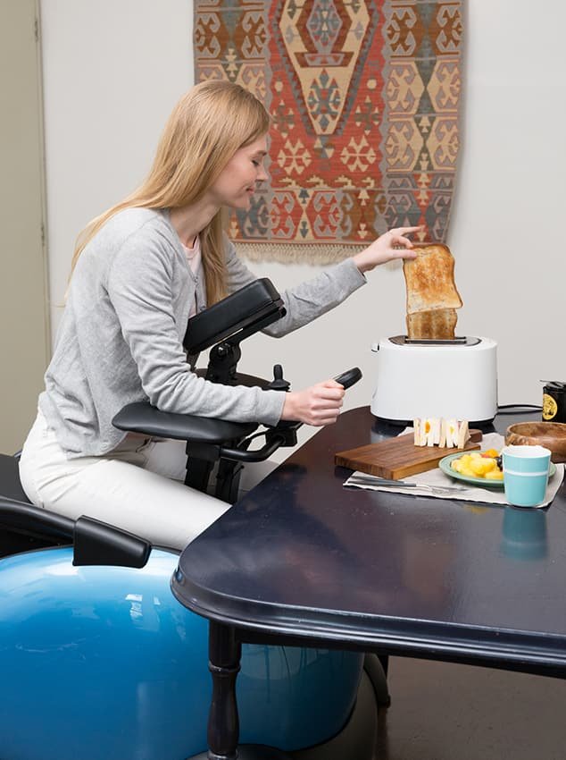 Descrição da imagem #PraTodosVerem: Imagem no formato retangular, na vertical. Uma mulher está sentada na cadeira de rodas robótica. Ela tem a parte da frente do tronco apoiado na cadeira, e está sentada em frente a uma mesa, comendo. A cadeira tem estrutura azul, rodas e acessórios pretos, além de formato oval. Fim da descrição.