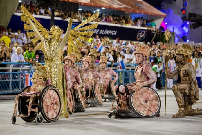 Descrição da imagem #PraTodosVerem: Imagem no formato retangular, na horizontal. Comissão de frente formada por cadeirantes, durante desfile do grupo especial no sambódromo do Rio de Janeiro. Fim da descrição.