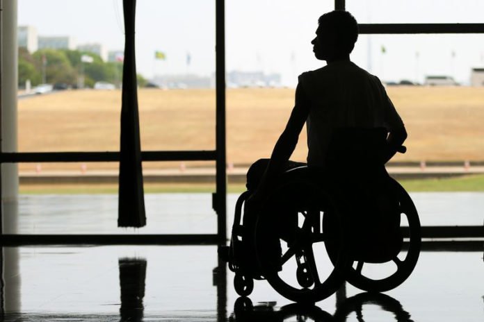 Descrição da imagem #PraTodosVerem: A imagem está no formato retangular, na horizontal. Um homem está em sua cadeira de rodas. A imagem foi tirada contra a luz e é possível ver apenas a sombra do homem, em frente a uma grande janela. Fim da descrição.